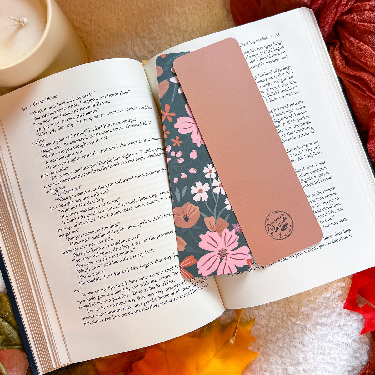 A rectangular rose gold bookmark with a floral pattern and a circular logo sits on the pages of an open book, styled with a candle, dried flowers, and autumn leaves.