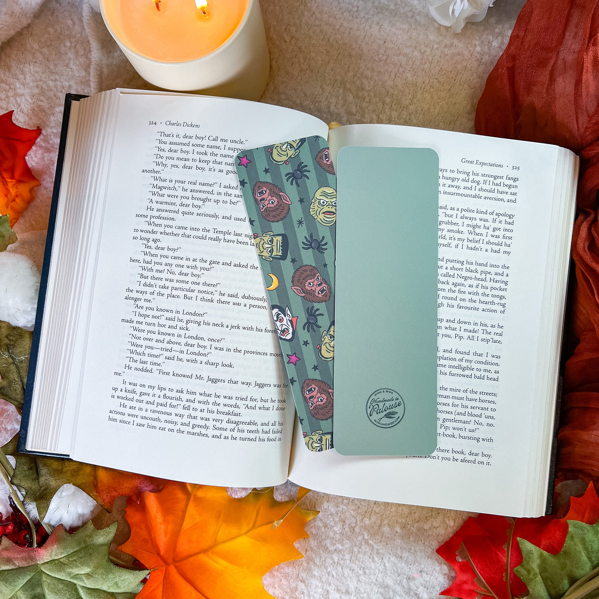 A decorative book bookmark with a teal background and colorful monster illustrations is shown inside an open book, styled with autumn leaves, a lit candle, and a cozy blanket.