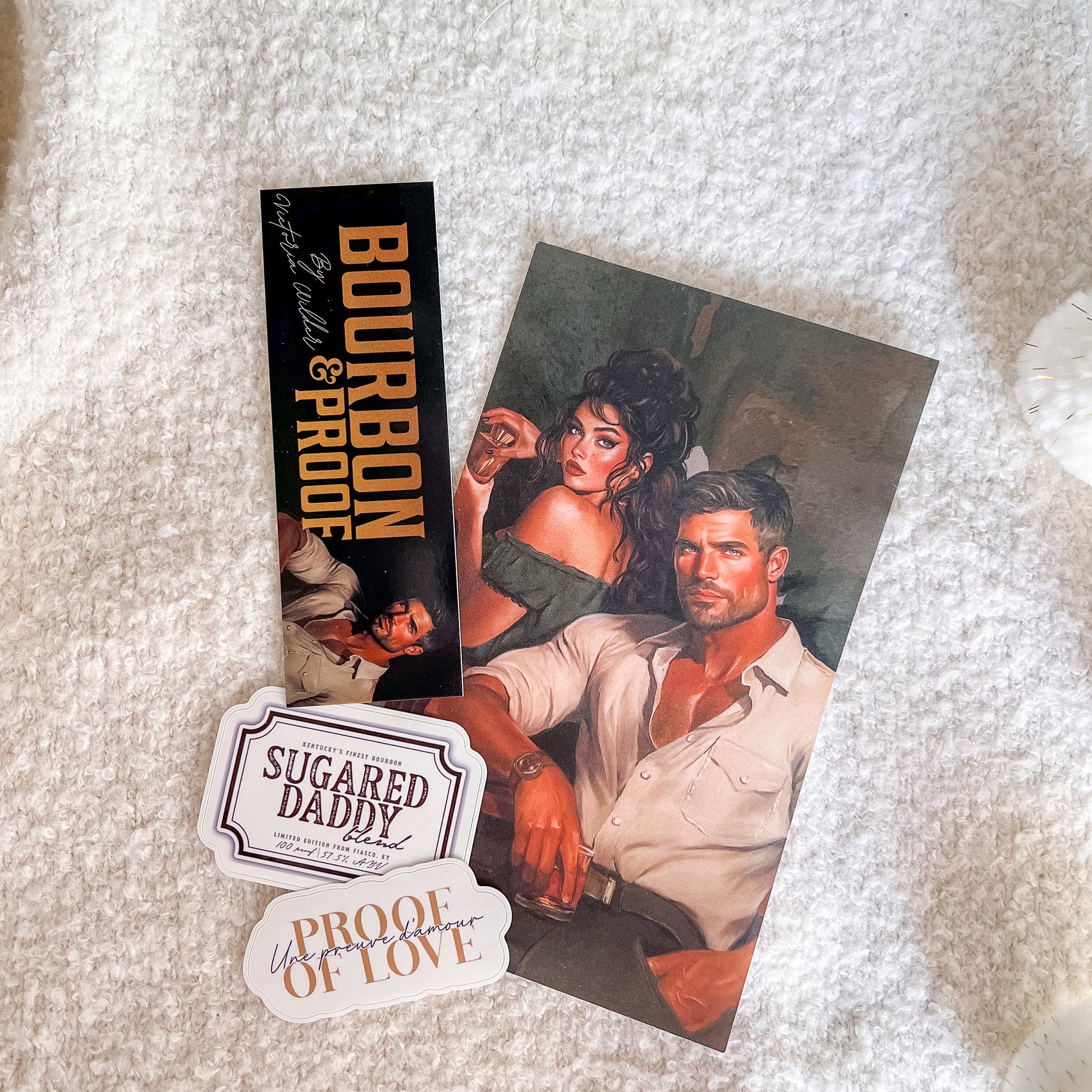 A flat-lay of printed merchandise on a white textured blanket, including a poster or art print with an illustrated couple, a black 'Bourbon & Proof' card, and two white stickers reading 'Sugared Daddy' and 'Proof of Love'.