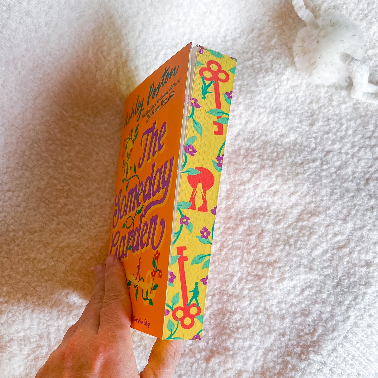 Colorful book titled 'The Someday Garden' held by a hand on a textured white surface.