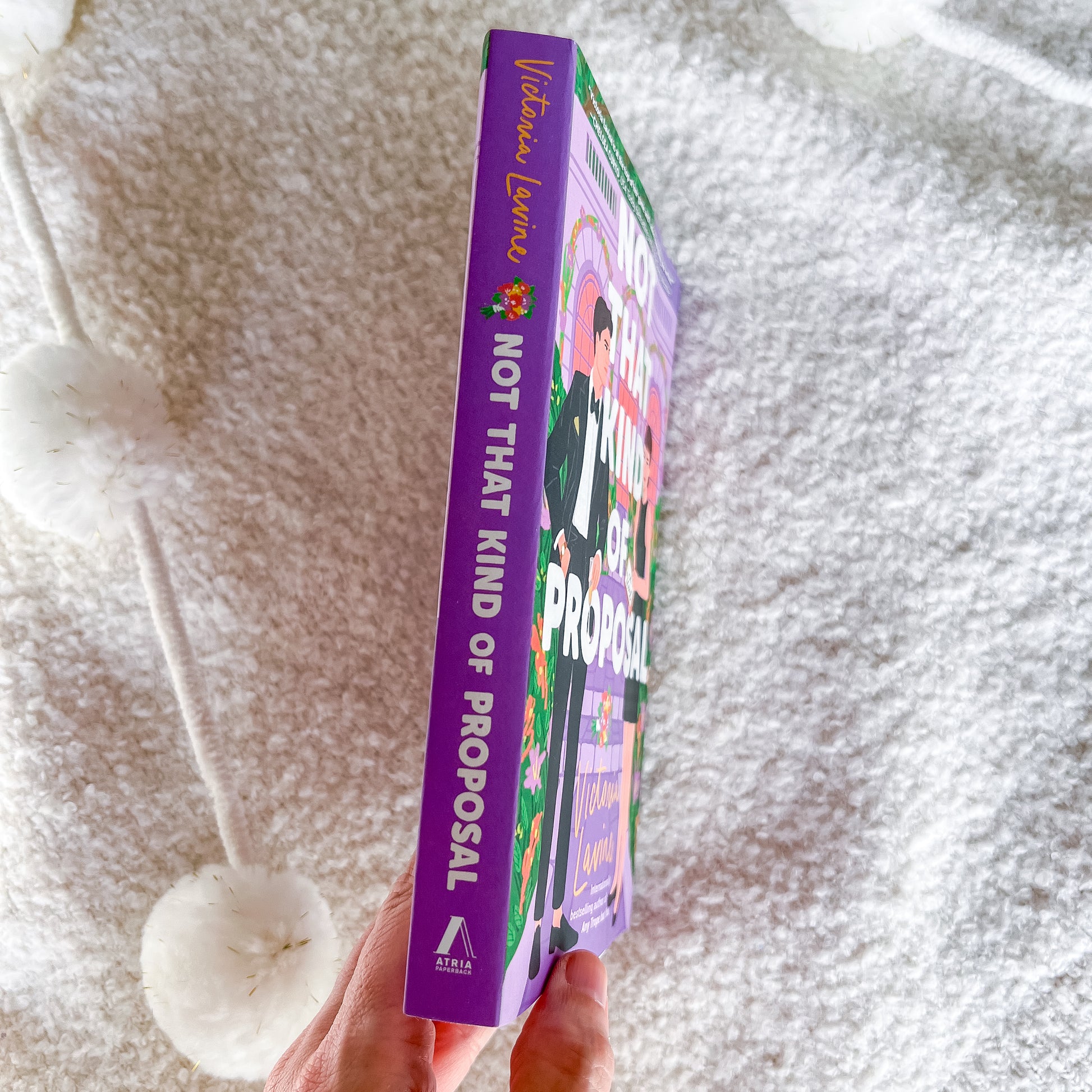 A hand holds a book titled 'NOT THAT KIND OF PROPOSAL' by Victoria Lundqvist. The book has a vibrant purple spine and cover with colorful illustrations, set against a soft, textured white blanket background.