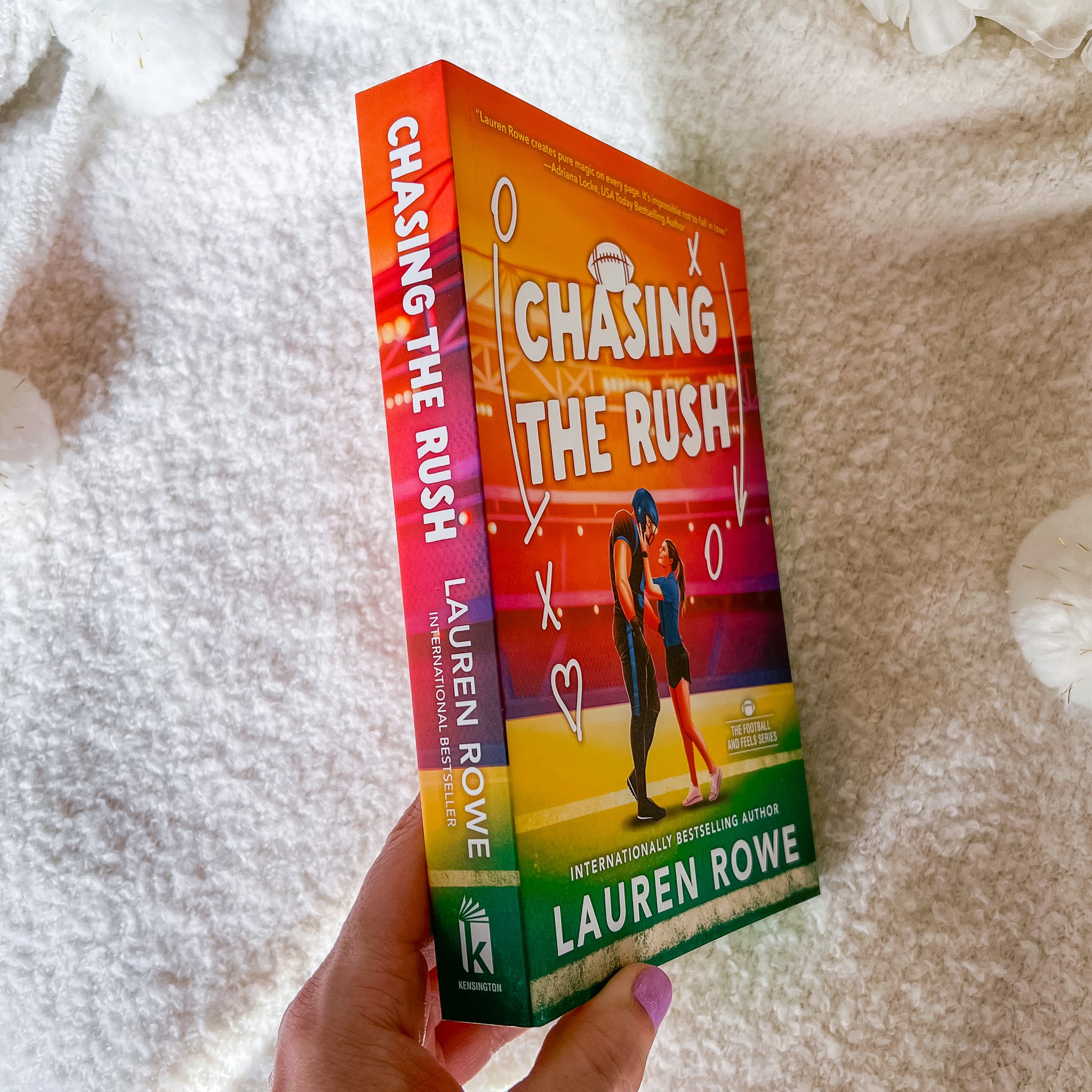 A hand holds the book 'Chasing the Rush' by Lauren Rowe, showing the colorful front cover and spine against a soft white textured blanket background.