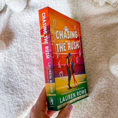 A hand holds the book 'Chasing the Rush' by Lauren Rowe, showing the colorful front cover and spine against a soft white textured blanket background.