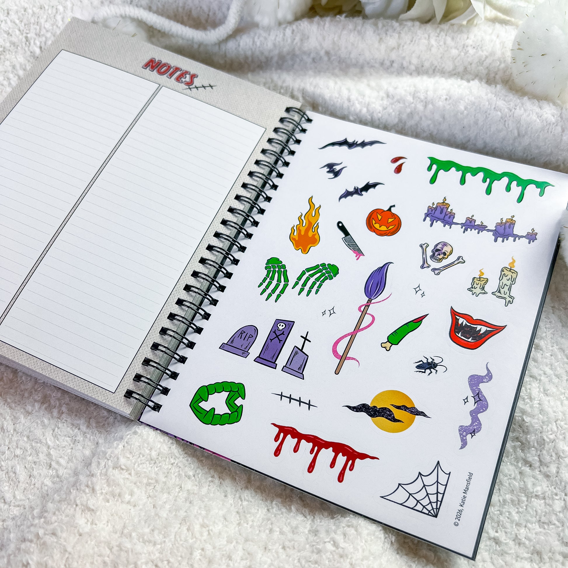 An open spiral notebook on a soft white blanket, showing a sheet of Halloween-themed temporary tattoos or stickers on the right page and lined notebook paper on the left.