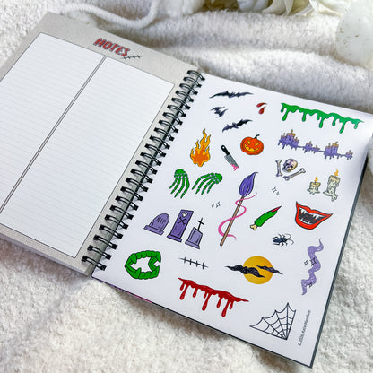 An open spiral notebook on a soft white blanket, showing a sheet of Halloween-themed temporary tattoos or stickers on the right page and lined notebook paper on the left.