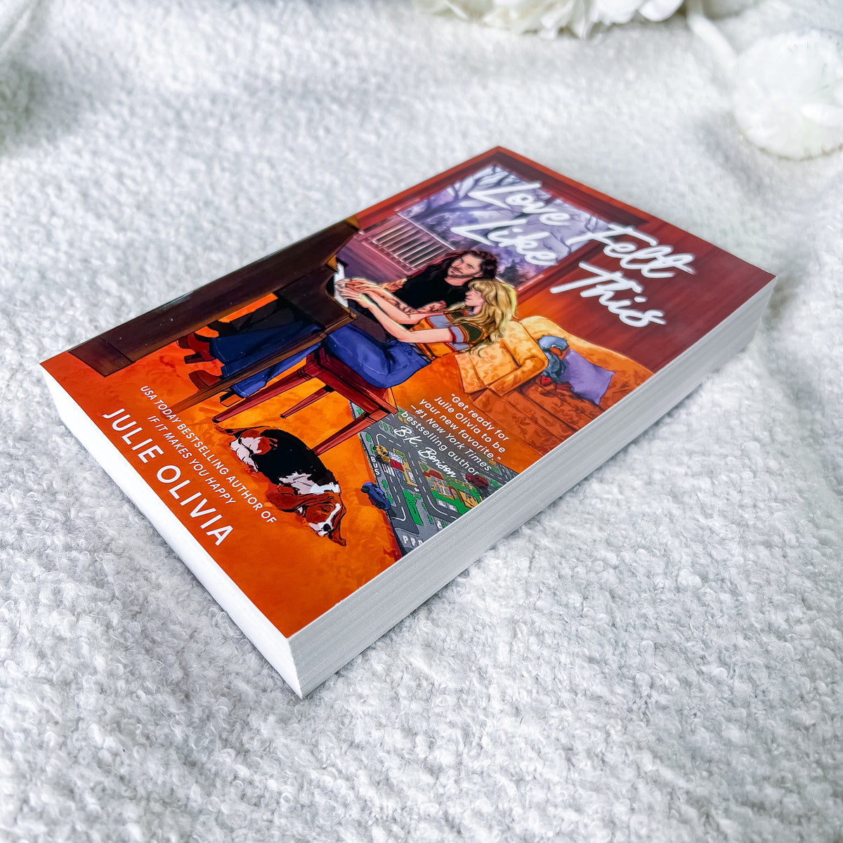 A paperback book titled 'What Happens When This Gets Too Hot' by Julie Olivia, featuring a vibrant orange and red cover with an illustration of a couple and a dog, resting on a soft white textured blanket.