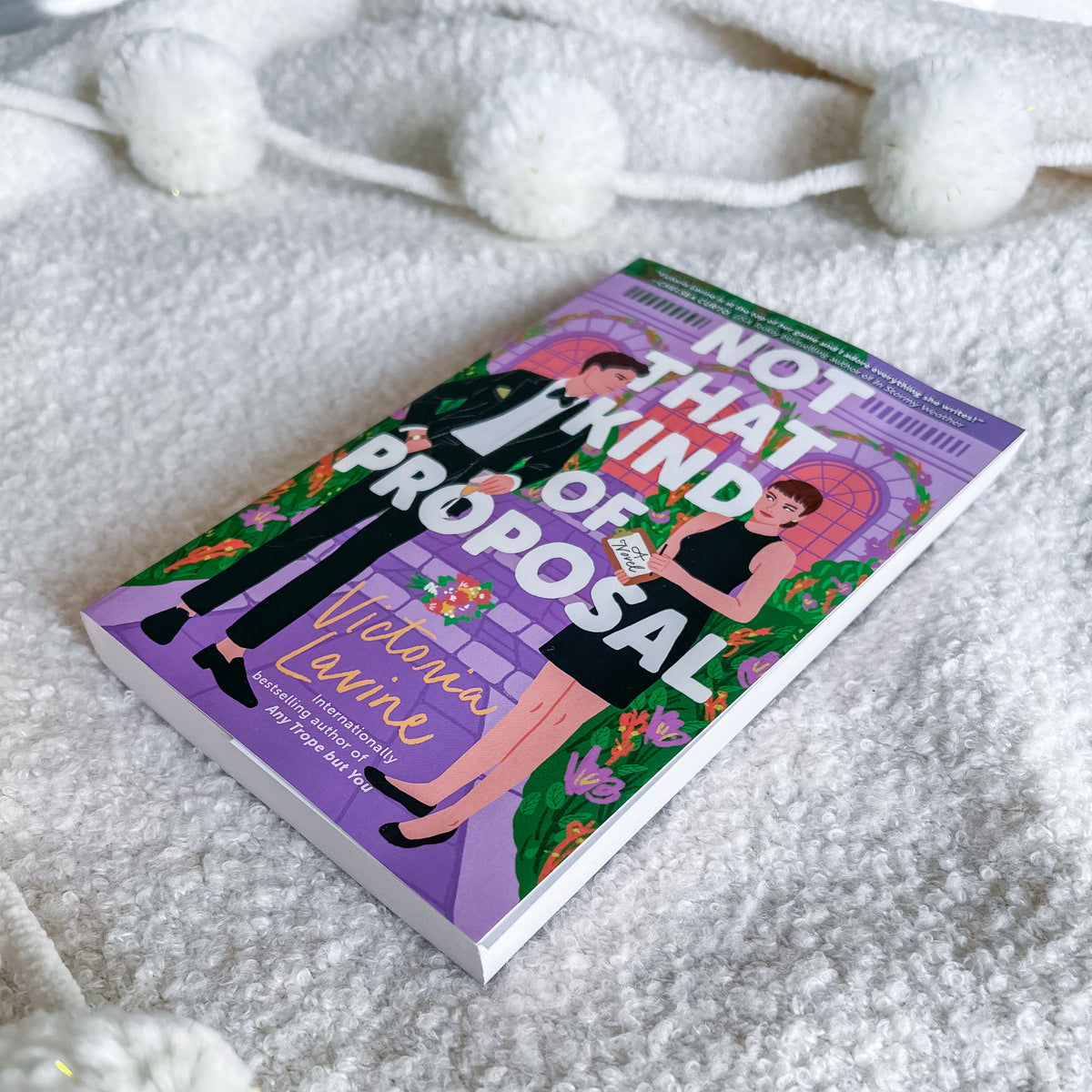 A book titled 'Not That Kind of Proposal' by Victoria Larrie lies on a white textured blanket with white pom-pom garlands in the background. The book cover features a colorful illustration of a couple with a wedding ring box.