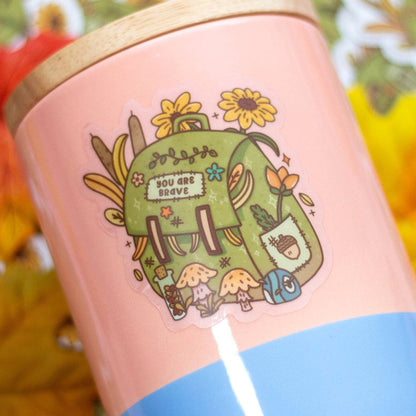 Pink mug with a green illustration of a backpack and flowers on a blurred autumn leaf background