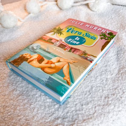 A hardcover book titled 'Vera Stein Is Fine' by Julie Murphy, featuring a colorful cover illustration of a woman by a pool, styled on a white textured blanket with white pom-pom garlands.