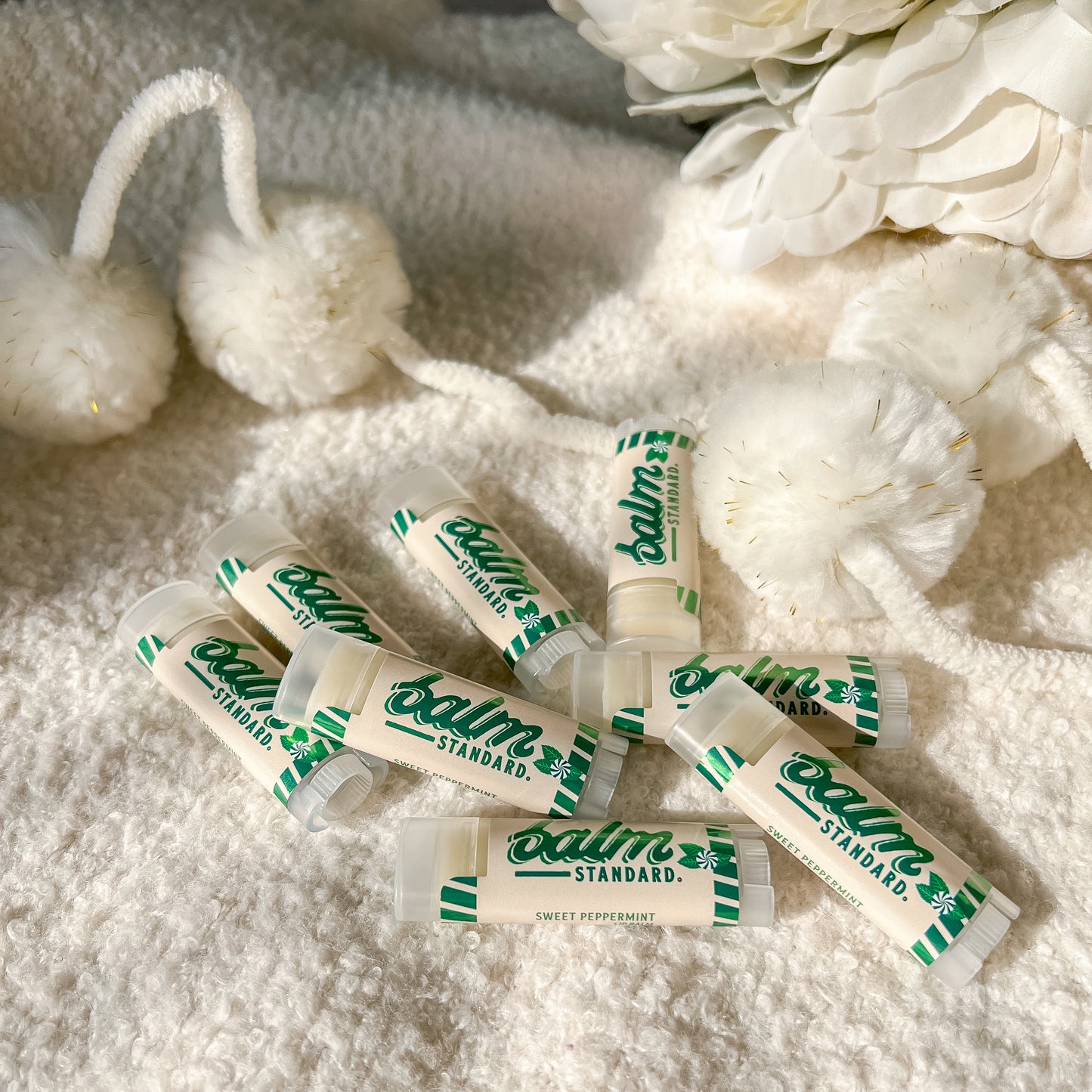 Six tubes of Balm Standard lip balm in Sweet Peppermint scent, arranged on a soft white textured blanket with decorative white flowers and pom-poms.