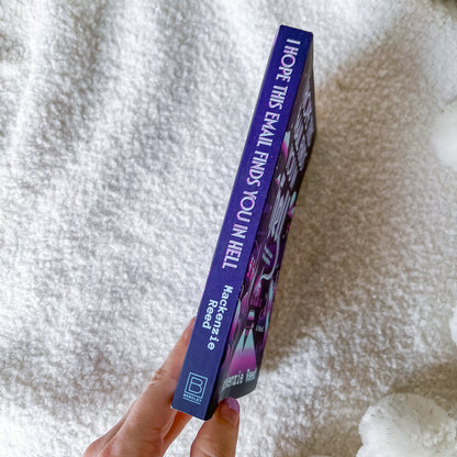 A hand holds a slim blue book titled 'I Hope This Email Finds You In Hell' by Meg Penrose, standing vertically against a soft white textured blanket background.