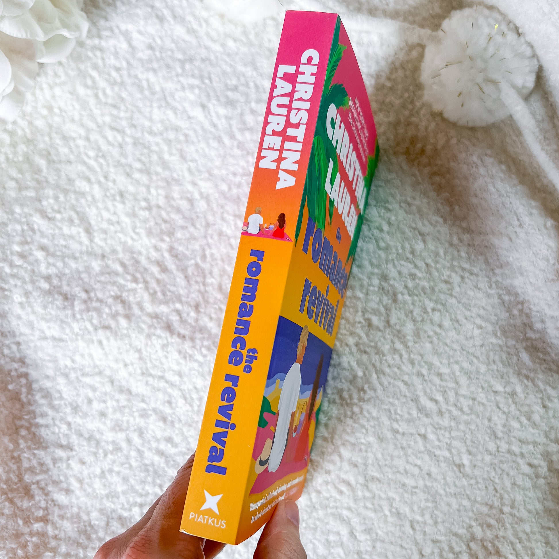 A hand holds a colorful paperback book titled 'Romance the Revival' by Christina Lauren. The book cover features a bright gradient of orange, pink, yellow, green, and blue with illustrations of a couple and a beach scene.
