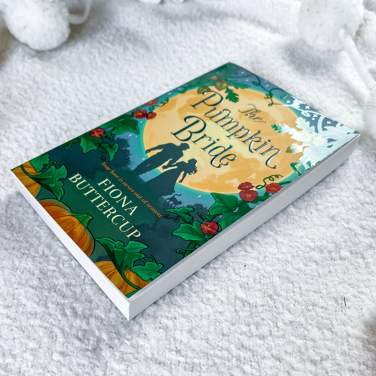 A hardcover book titled "The Pumpkin Bride" by Fiona Buttercup lies on a soft white textured blanket. The cover features a detailed autumn illustration with a large yellow pumpkin, orange pumpkins, green leaves, red berries, and a silhouette of a bride and groom.