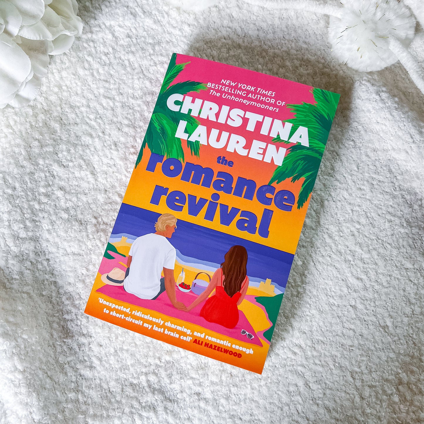 The book cover of 'The Romance Revival' by Christina Lauren is displayed diagonally on a textured white blanket with white flowers in the corners. The cover features a colorful illustration of a couple on a beach with palm trees and a gradient background of pink, orange, and blue.