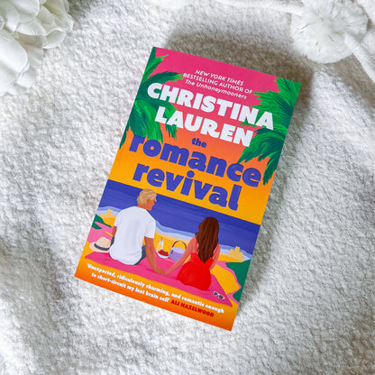 The book cover of 'The Romance Revival' by Christina Lauren is displayed diagonally on a textured white blanket with white flowers in the corners. The cover features a colorful illustration of a couple on a beach with palm trees and a gradient background of pink, orange, and blue.