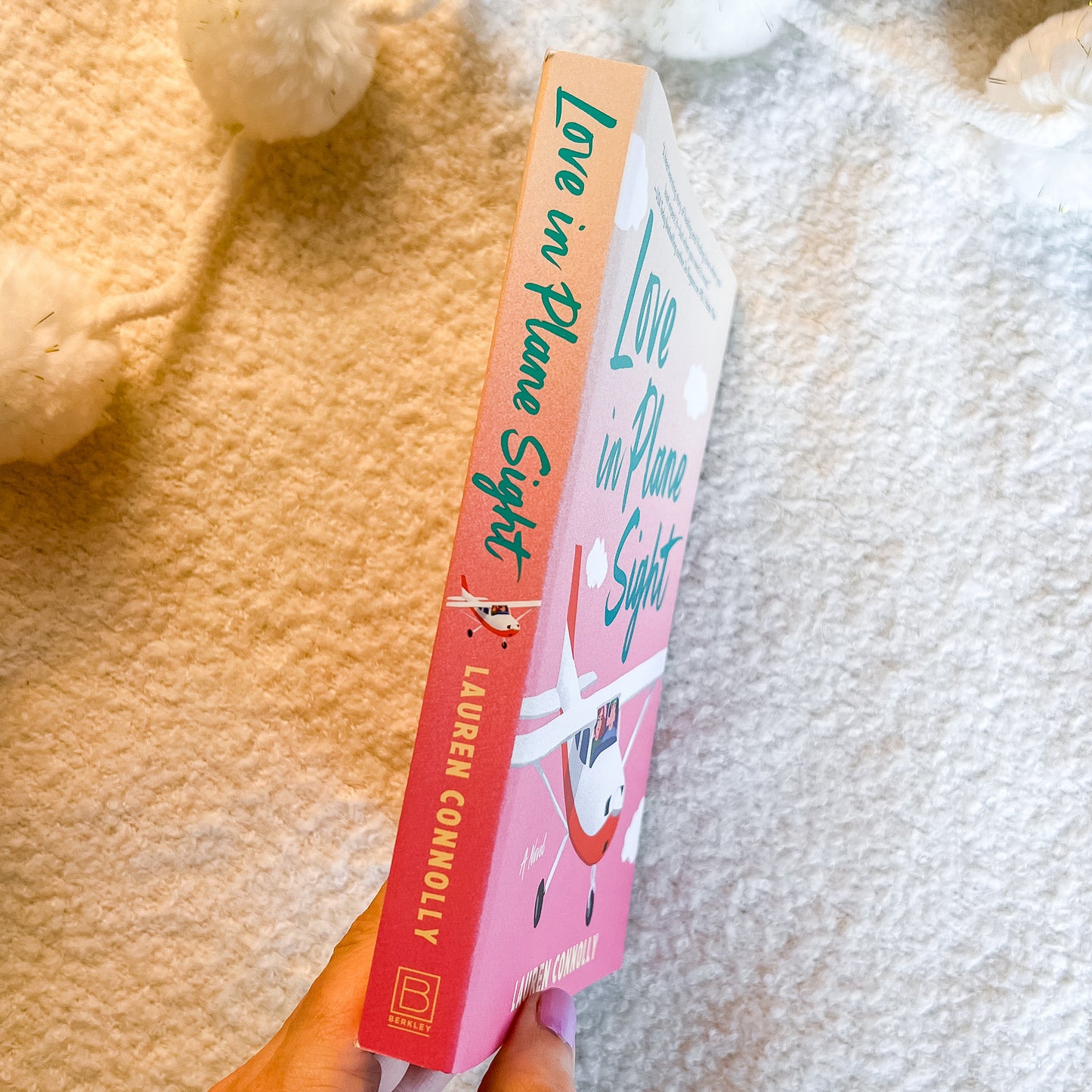 A hand holds a book titled 'Love in Plane Sight' by Lauren Connolly. The book cover features a pink and peach gradient with a cartoon rabbit illustration and teal lettering.