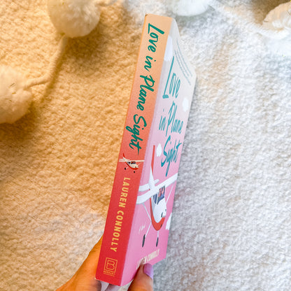 A hand holds a book titled 'Love in Plane Sight' by Lauren Connolly. The book cover features a pink and peach gradient with a cartoon rabbit illustration and teal lettering.