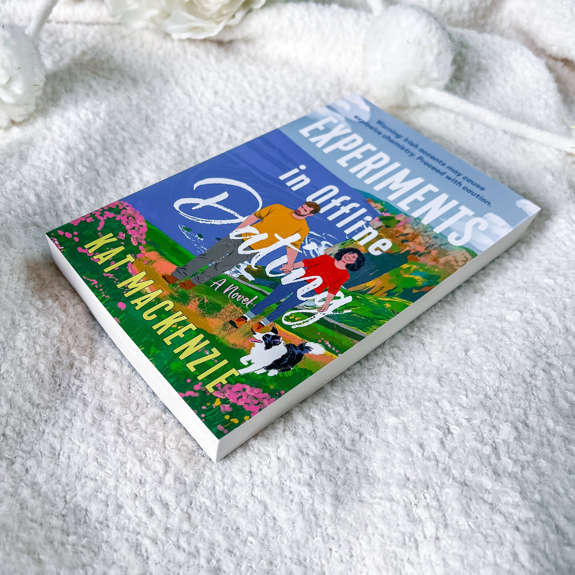 A book titled "Experiments in Offline Dating" by Jay MacKenzie lies diagonally on a soft white textured blanket, with white flowers blurred in the background.
