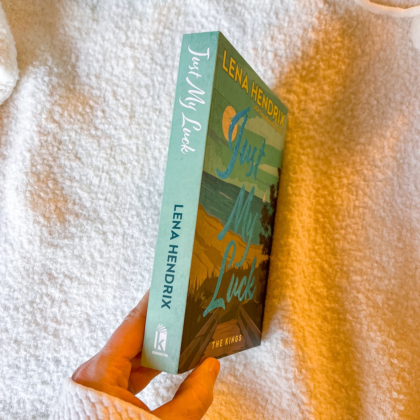 Book titled 'Just My Luck' by Lena Hendrix held against a textured white and orange background