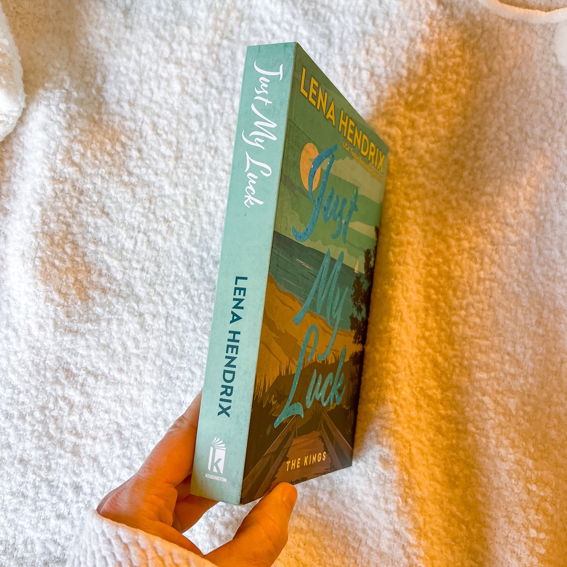 Book titled 'Just My Luck' by Lena Hendrix held against a textured white and orange background