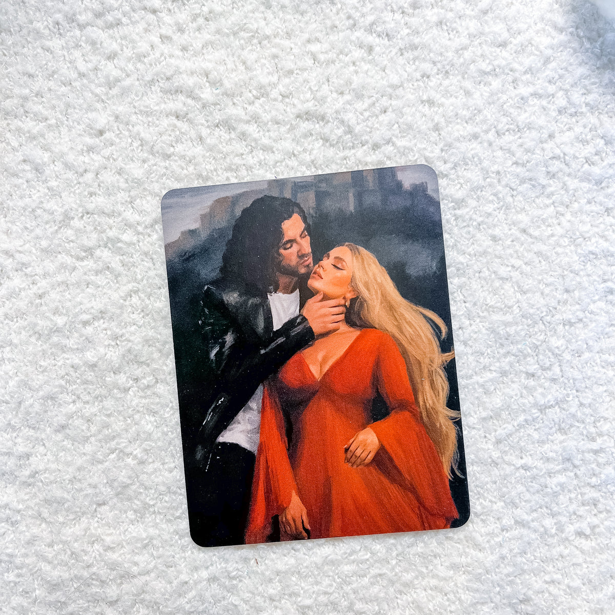 A rectangular art print or card featuring a painted couple embracing, placed on a textured white fabric background.