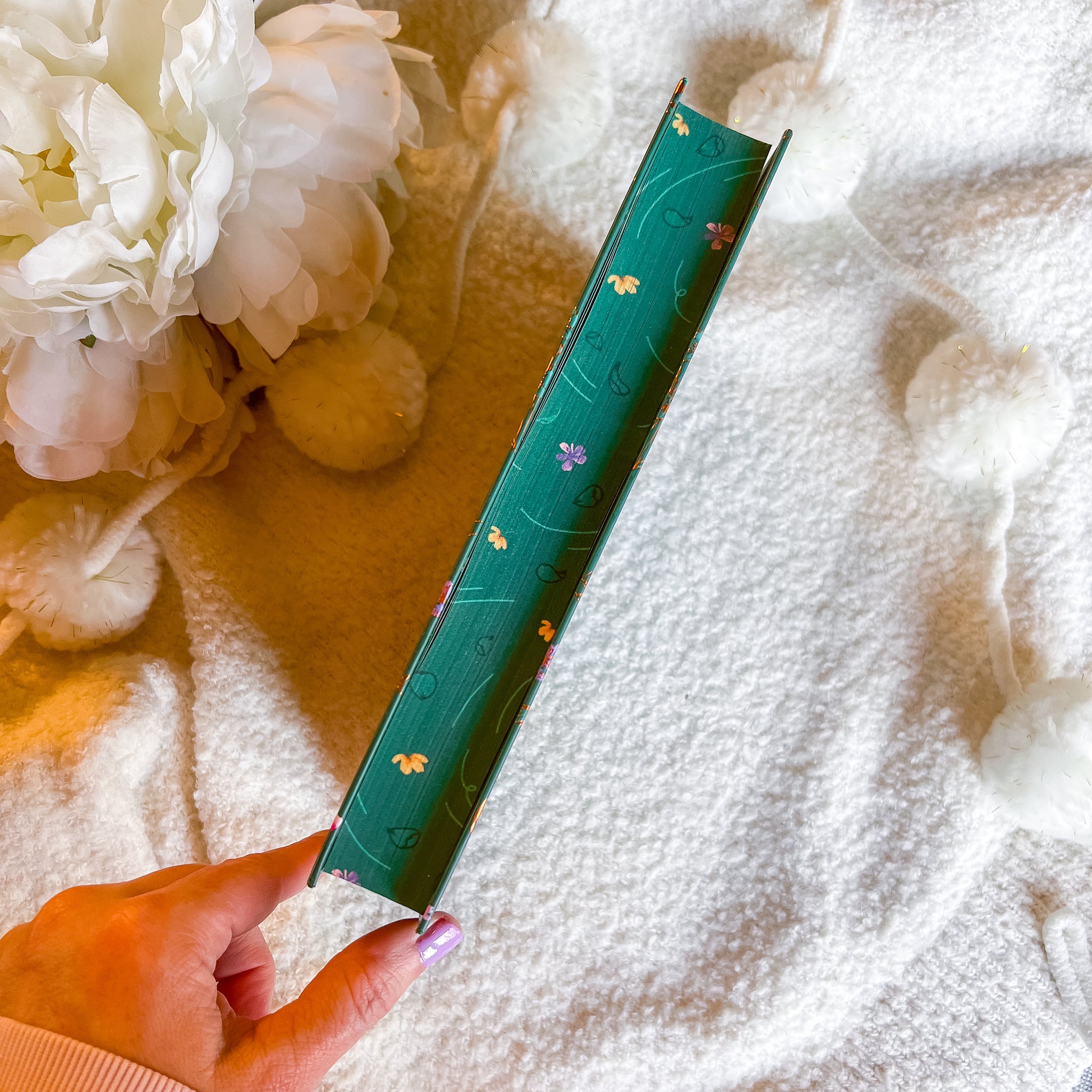 Green book with floral design held by a hand on a textured surface with flowers in the background