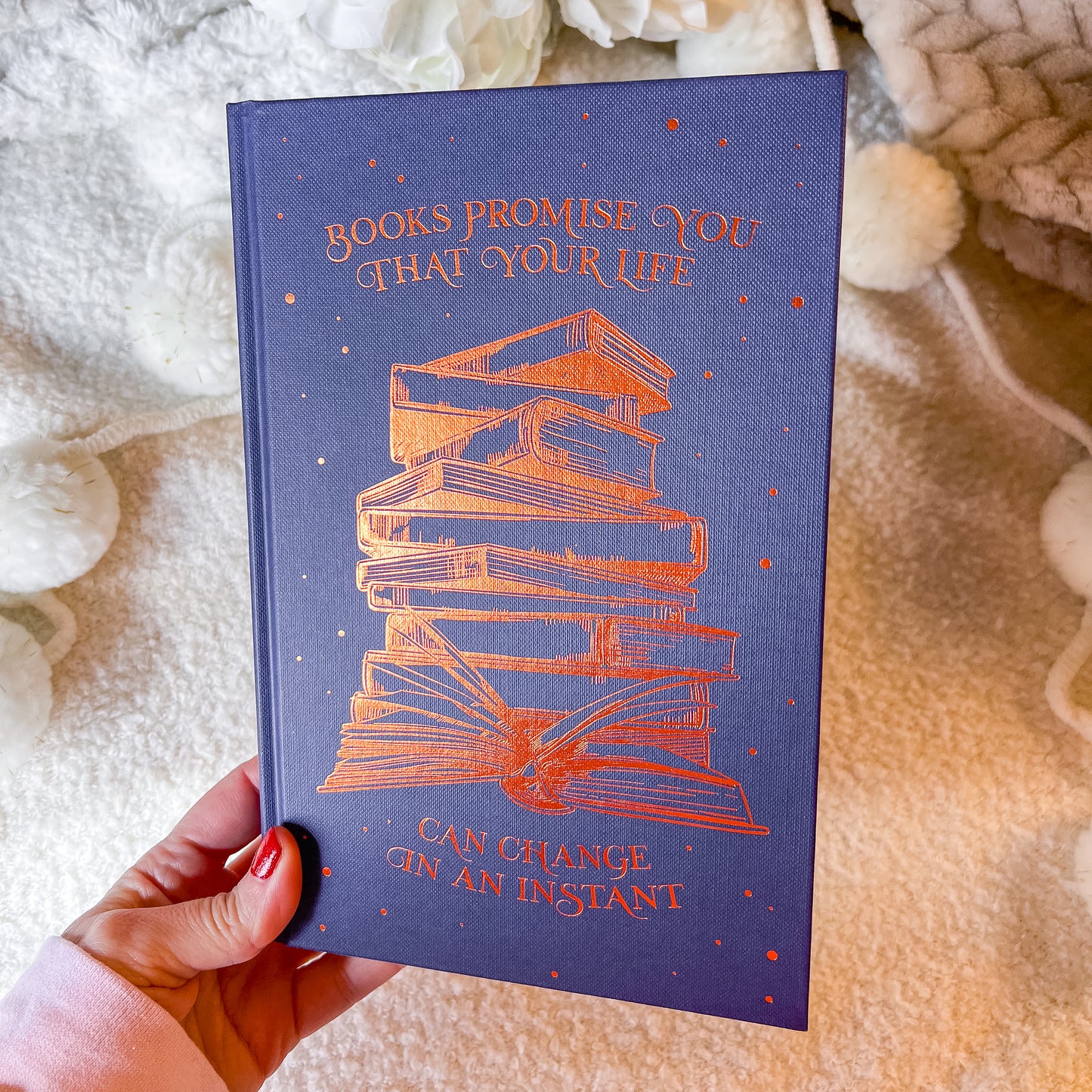 Book with a stack of books illustration and text on a blue background, held against a textured fabric background.