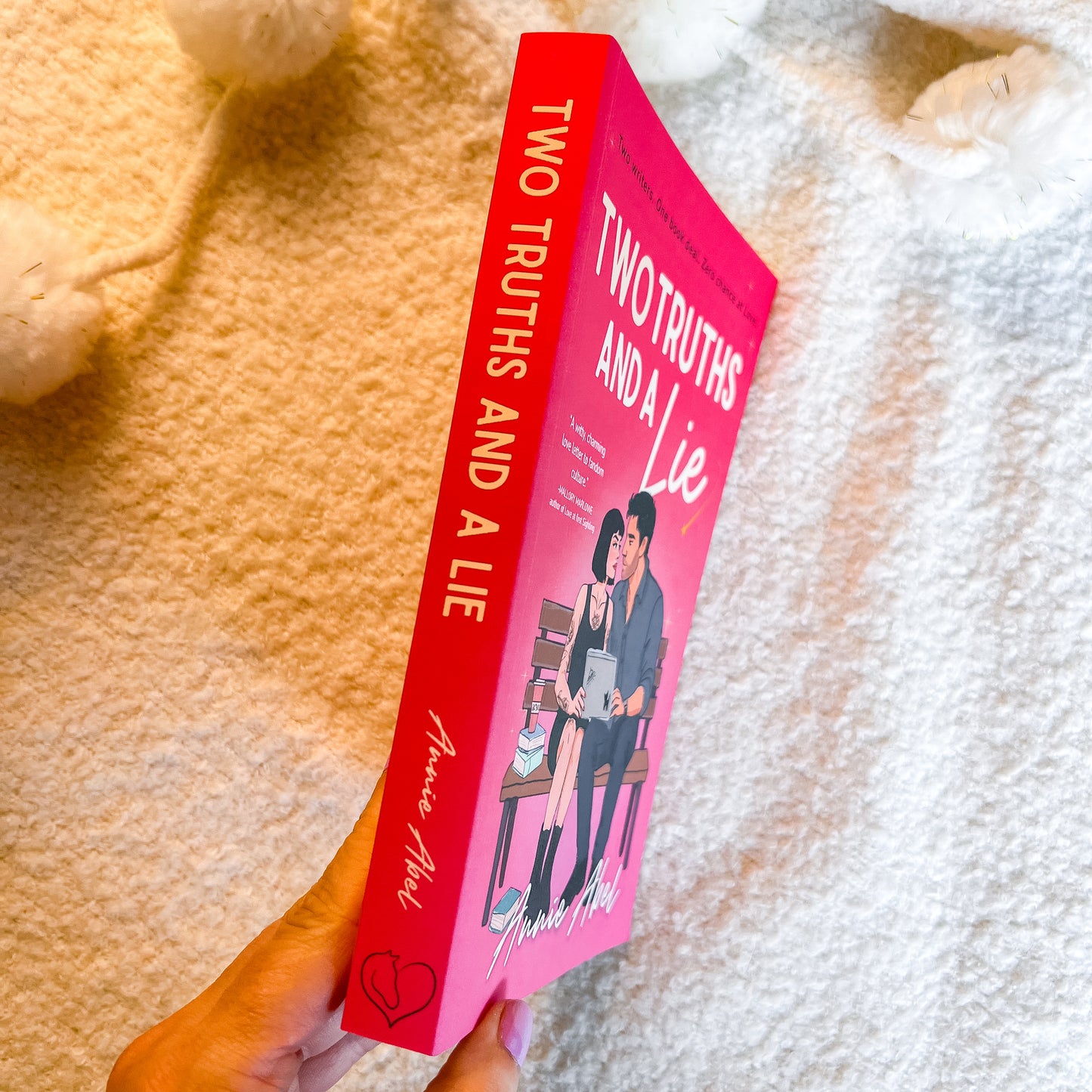 A hand holds a pink romance novel titled 'Two Truths and a Lie' by against a soft, textured cream blanket background.