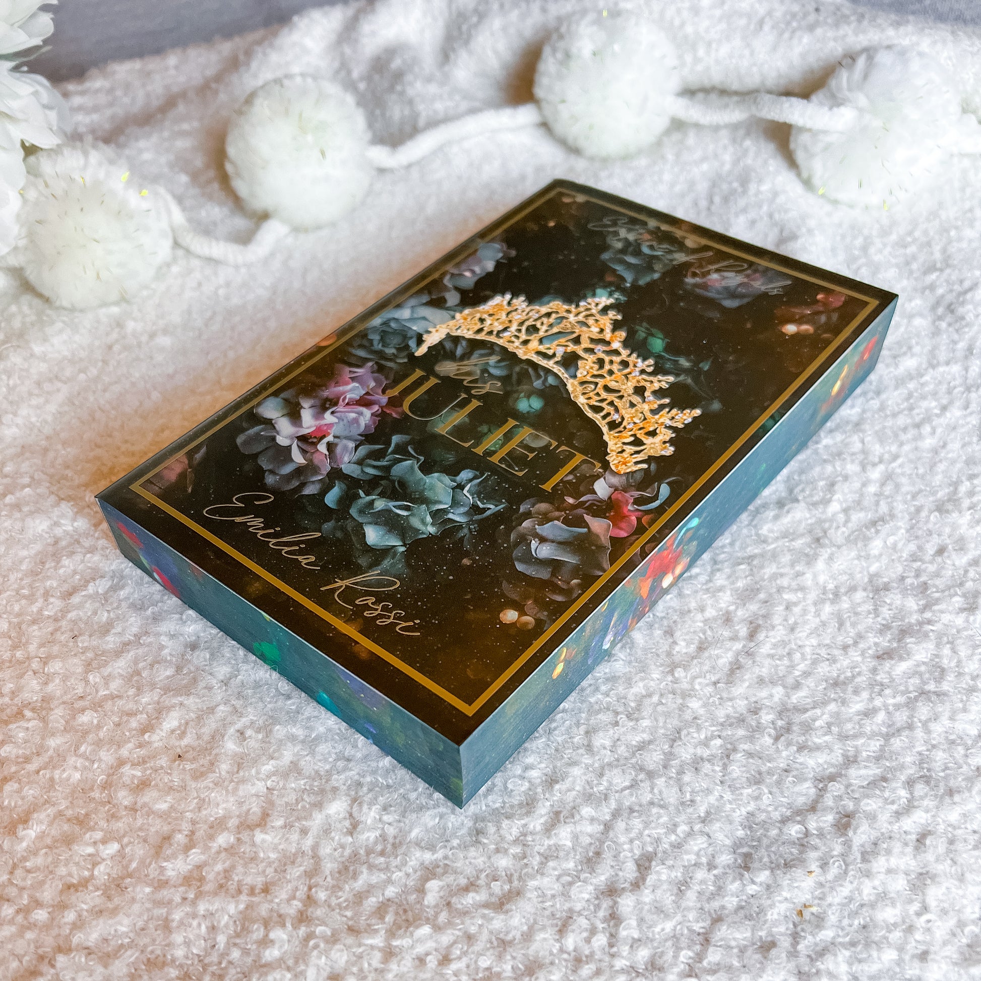 A decorative rectangular box labeled 'Juliet' and 'Emilie Rose' lies on a soft white textured blanket, with white pom-pom garlands blurred in the background. The box has a dark floral design with gold lettering and a gold crown motif on top.