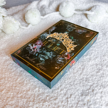 A decorative rectangular box labeled 'Juliet' and 'Emilie Rose' lies on a soft white textured blanket, with white pom-pom garlands blurred in the background. The box has a dark floral design with gold lettering and a gold crown motif on top.