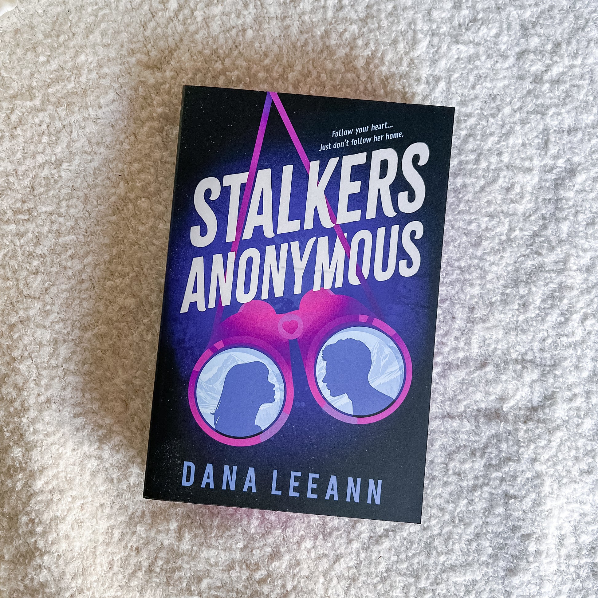 The book cover of 'Stalkers Anonymous' by Dana Leeann, featuring a dark purple and black background with a pink binocular graphic and silhouettes, photographed on a textured beige carpet.