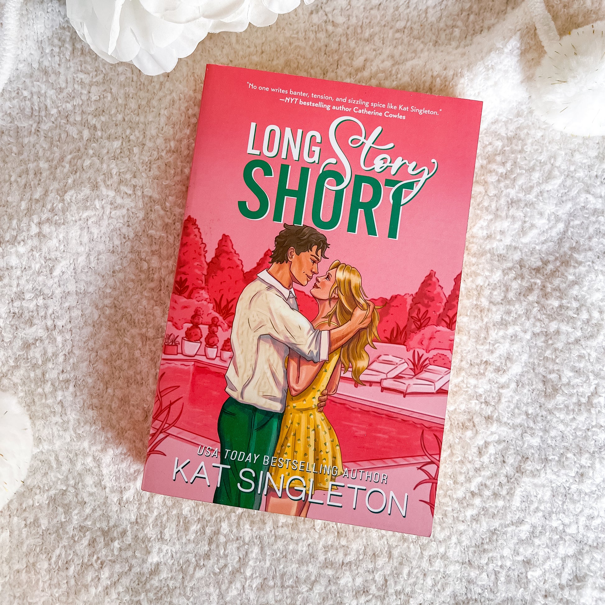 A book titled "Long Story Short" by Kat Singleton lies on a soft white textured blanket. The cover features a pink-toned illustration of a couple embracing, with the title in large white and green lettering.