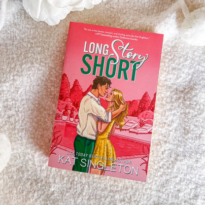 A book titled "Long Story Short" by Kat Singleton lies on a soft white textured blanket. The cover features a pink-toned illustration of a couple embracing, with the title in large white and green lettering.