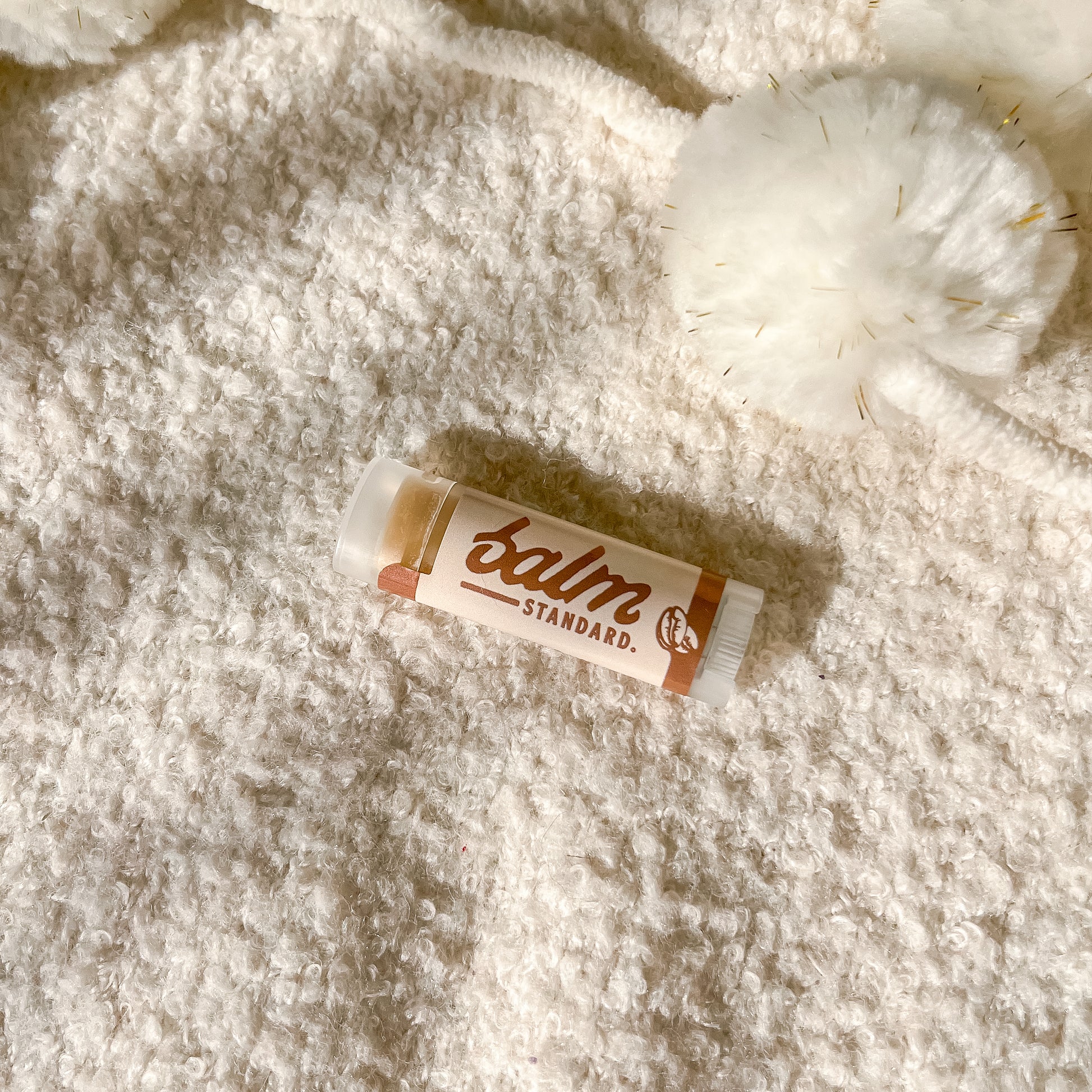 A tube of lip balm labeled 'balm STANDARD.' lies on a soft white textured blanket with a fluffy pom-pom nearby.