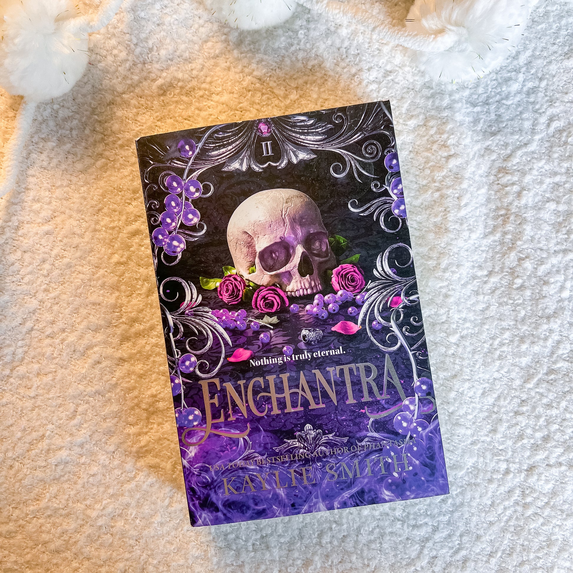 The book is displayed diagonally on a soft, textured cream-colored blanket, surrounded by decorative white flowers in the corners. The cover features a dark, gothic design with a central skull, purple roses, and ornate silver filigree.
