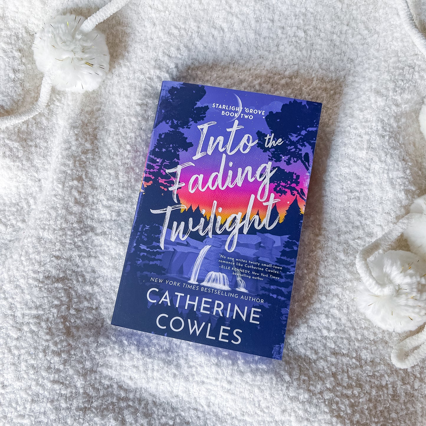 A book titled "Into the Fading Twilight" by Catherine Cowles lies on a soft white textured blanket, surrounded by white decorative pom-pom ornaments.