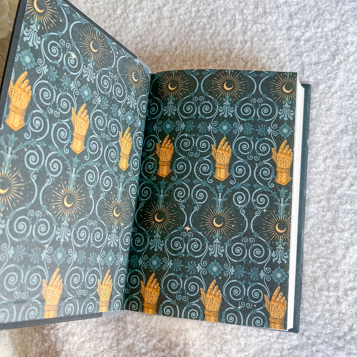 An open book laid on a light gray textured surface, showing an ornate repeating pattern with gold-toned hands, crescent moons, suns, and swirling decorative motifs on a dark teal background.