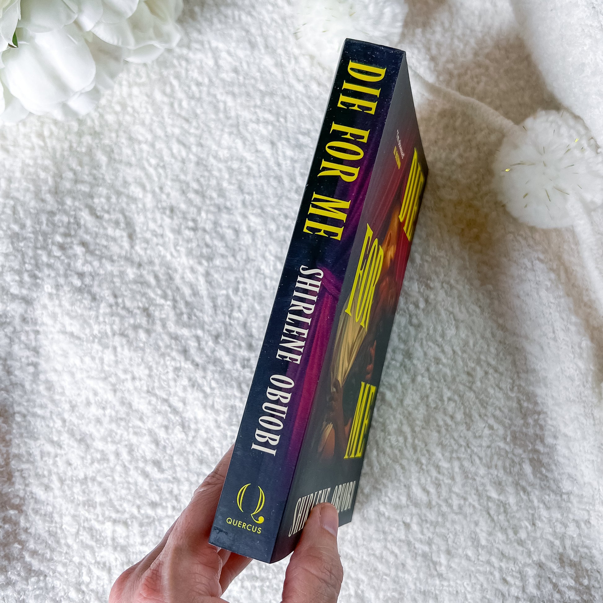 A hand holds a paperback book titled 'Die for Me' by Shirlene, against a soft white textured blanket background with white flowers in the corner.