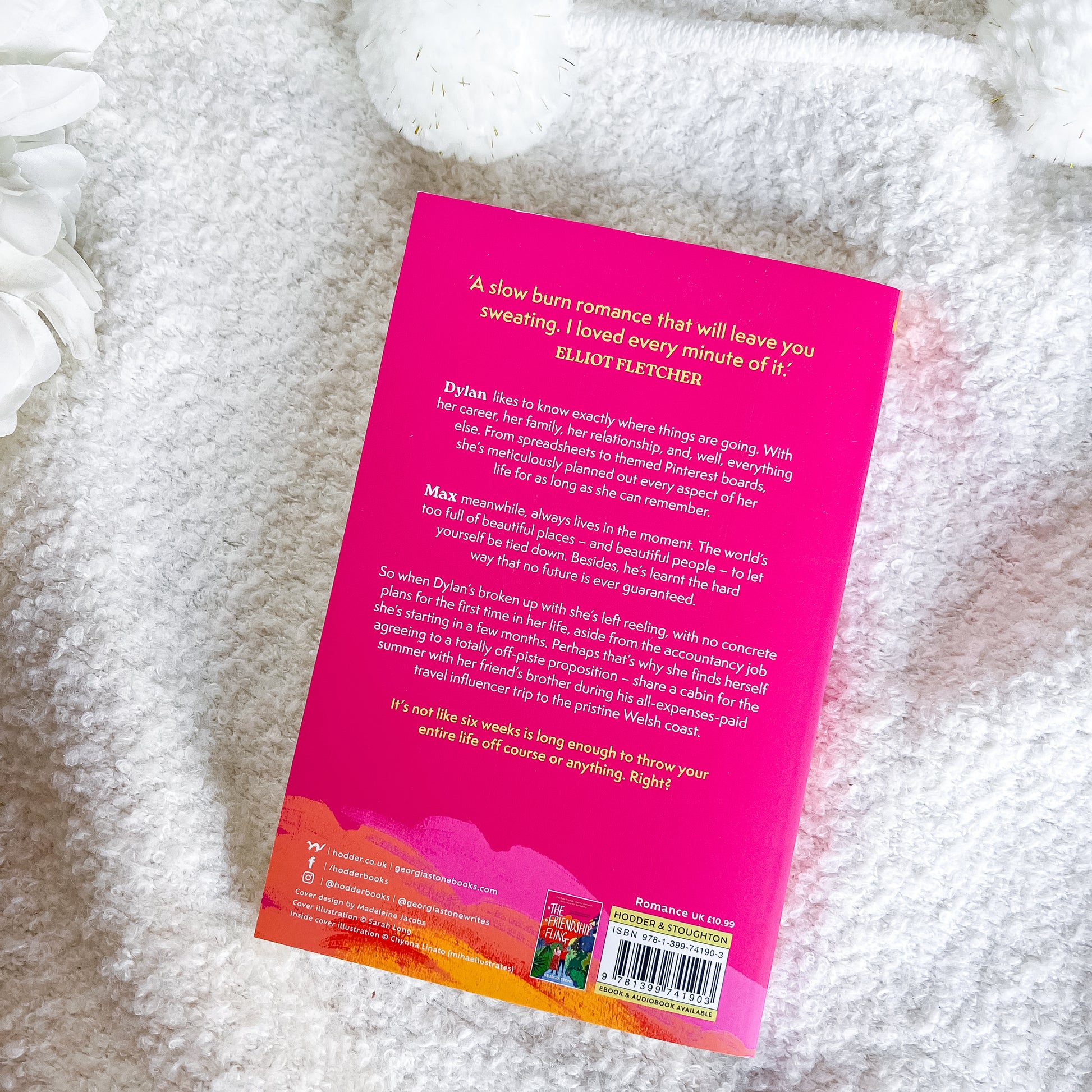 The back cover of a book lies flat on a soft, white textured blanket. The cover features a vibrant hot pink background with white text, a colorful orange and yellow abstract wave design at the bottom, and a barcode on the lower right.
