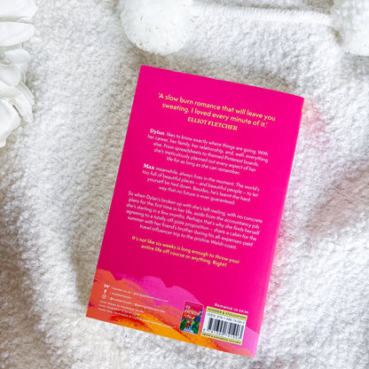 The back cover of a book lies flat on a soft, white textured blanket. The cover features a vibrant hot pink background with white text, a colorful orange and yellow abstract wave design at the bottom, and a barcode on the lower right.