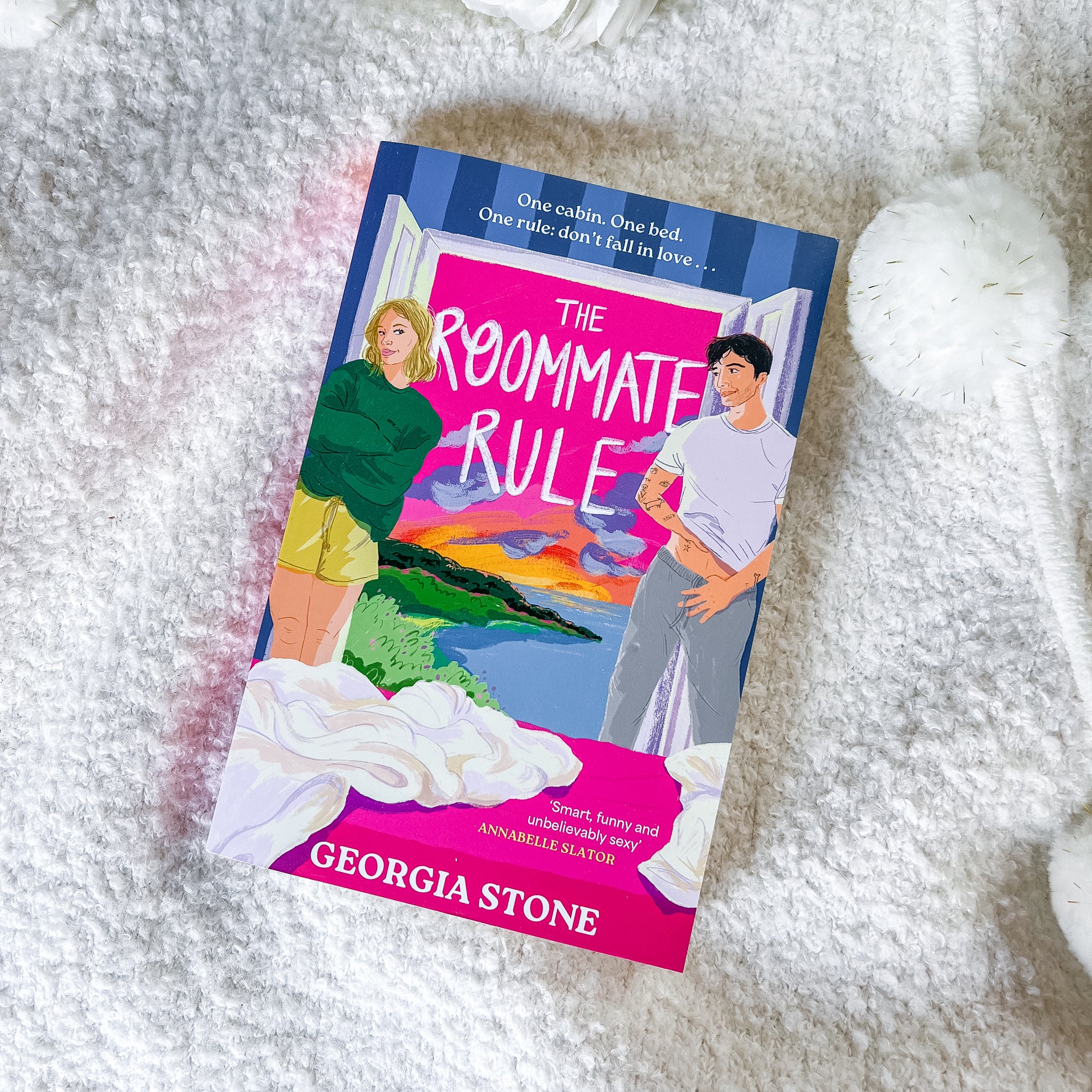 The book 'The Roommate Rule' by Georgia Stone is shown lying on a white textured blanket, with a white pom-pom toy and a glass of water nearby.
