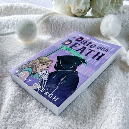 A paperback book titled "A Date with Death" by Kelly Creagh lies on a soft white textured blanket, styled with white flowers and a white pom-pom in the background.