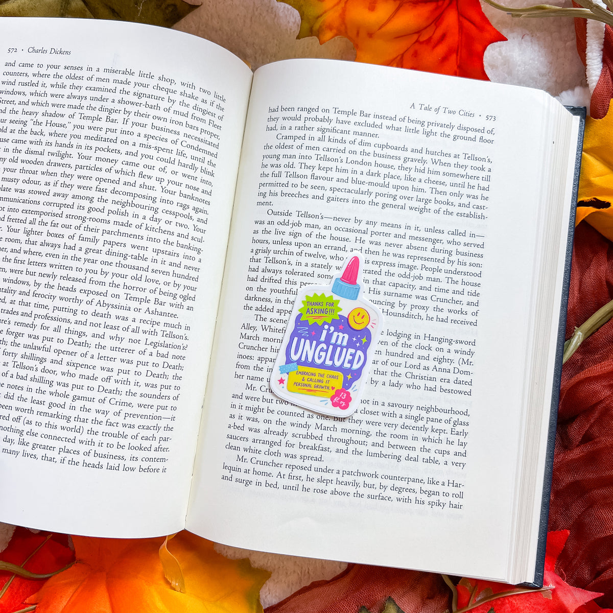 An open book spread showing printed text pages, with a colorful 'I'm in UNGUILED' sticker placed over a section. Red rose petals are scattered around the open book on a dark surface.