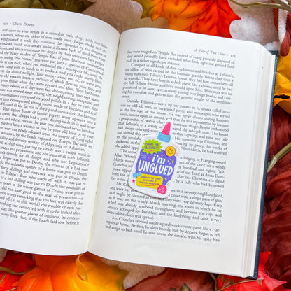 An open book spread showing printed text pages, with a colorful 'I'm in UNGUILED' sticker placed over a section. Red rose petals are scattered around the open book on a dark surface.