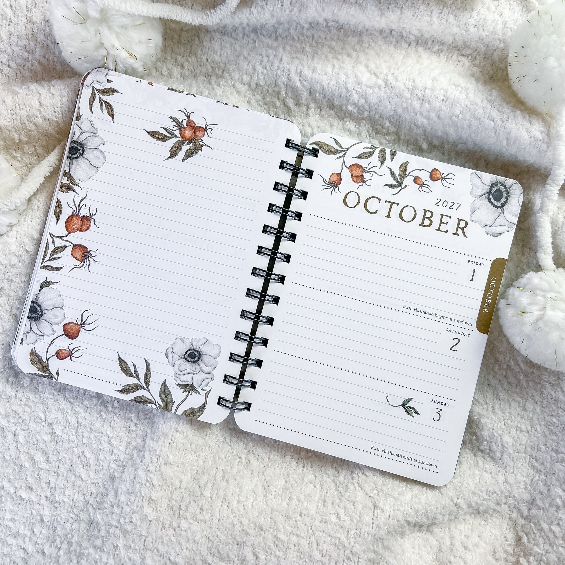 An open spiral-bound planner or calendar showing October 2027, laid on a soft cream knit blanket with white pom-pom accents. The pages have a floral and botanical border print with white flowers, gray leaves, and small orange berries, plus a gold vertical accent near the right edge.