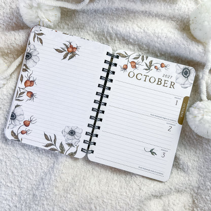 An open spiral-bound planner or calendar showing October 2027, laid on a soft cream knit blanket with white pom-pom accents. The pages have a floral and botanical border print with white flowers, gray leaves, and small orange berries, plus a gold vertical accent near the right edge.