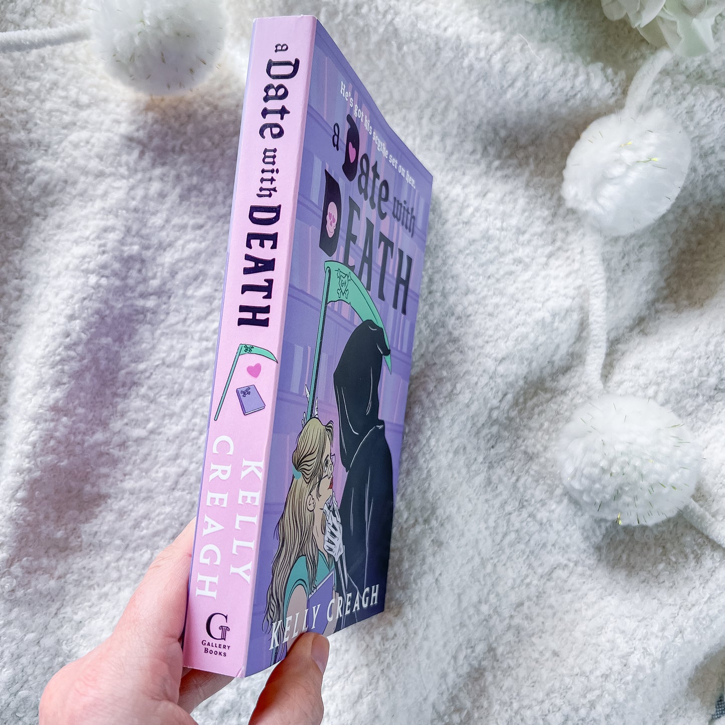 A hand holds a book titled 'A Date with Death' by Kelly Creagh. The book has a lavender-purple cover and spine with black and teal text and illustration of a hooded figure holding a scythe.