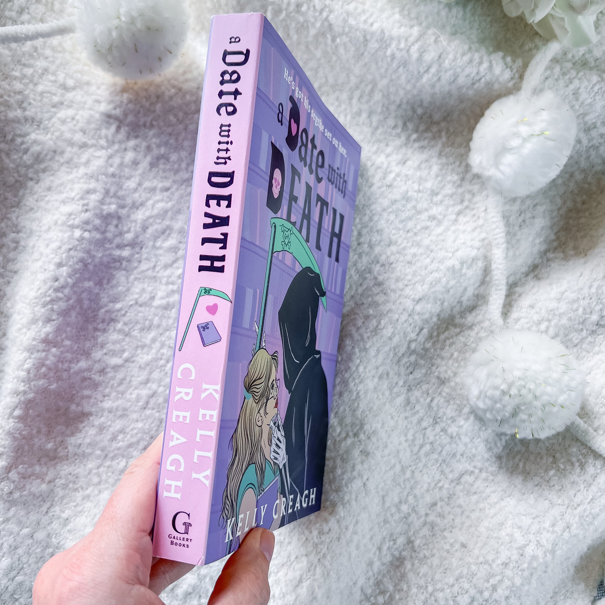 A hand holds a book titled 'A Date with Death' by Kelly Creagh. The book has a lavender-purple cover and spine with black and teal text and illustration of a hooded figure holding a scythe.
