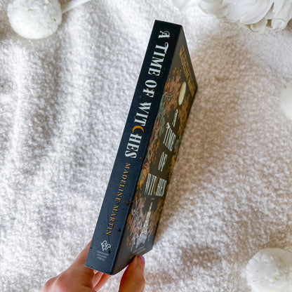A hand holds a copy of the book 'A Time of Witches' by Madeleine Martin, showing the dark blue spine with gold and white lettering, set against a soft white textured blanket with decorative white flowers.