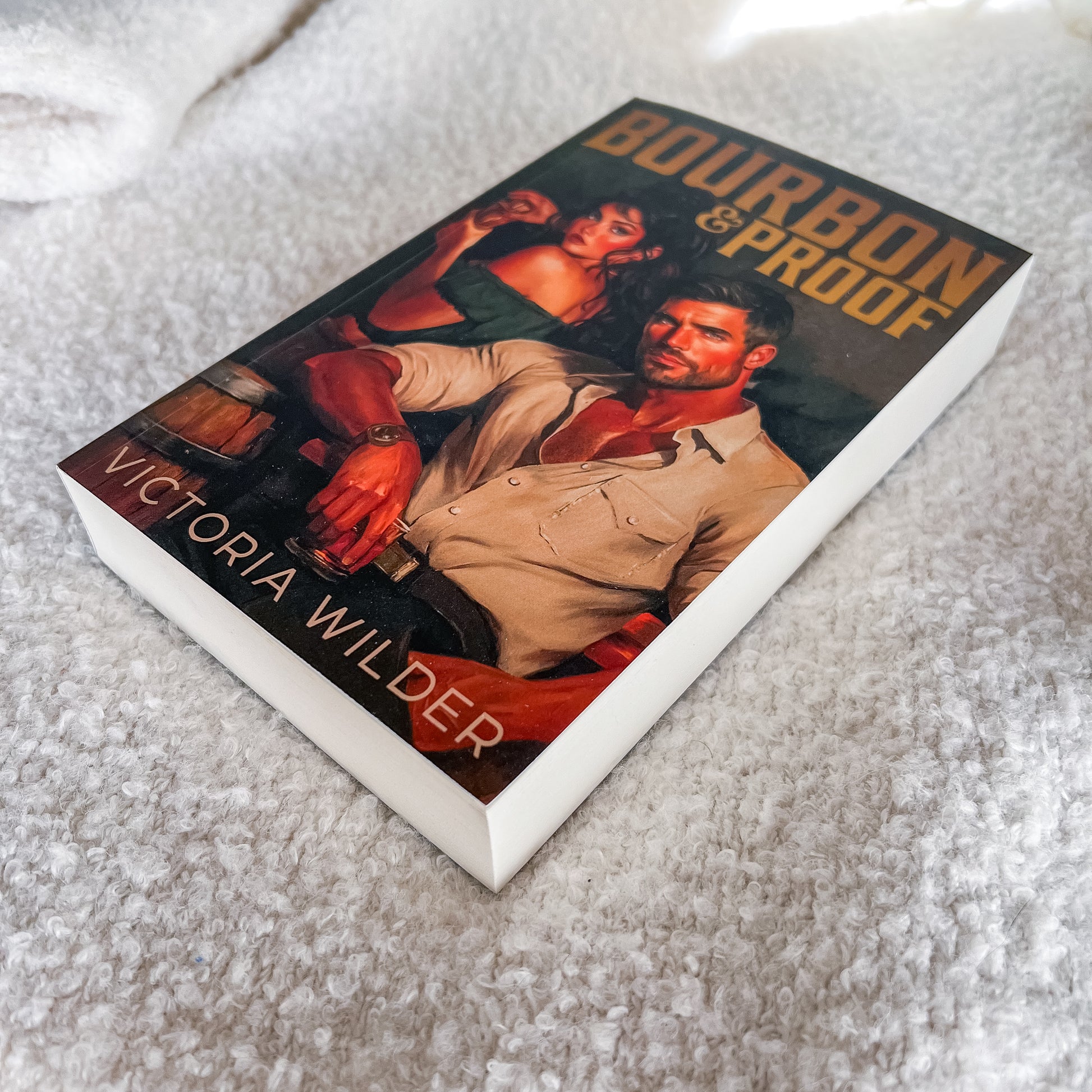 A paperback copy of the book 'Bourbon & Proof' by Victoria Wilder, featuring a romantic cover illustration of a man and woman on a bed with white textured sheets.